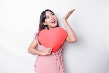 Portrait of a smiling Asian woman holding a big red heart symbol pointing up at copy space isolated over white background