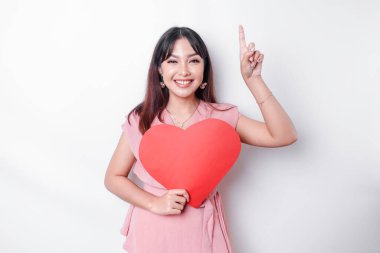 Portrait of a smiling Asian woman holding a big red heart symbol pointing up at copy space isolated over white background