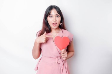A shocked Asian woman dressed in pink, pointing at the copy space beside her while holding a big red heart-shaped paper, isolated by a white background. Valentine's Day concept.