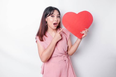 A shocked Asian woman dressed in pink, pointing at the copy space beside her while holding a big red heart-shaped paper, isolated by a white background. Valentine's Day concept.
