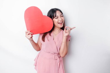 Excited Asian woman dressed in pink, pointing at the copy space beside her while holding a big red heart-shaped paper, isolated by white background