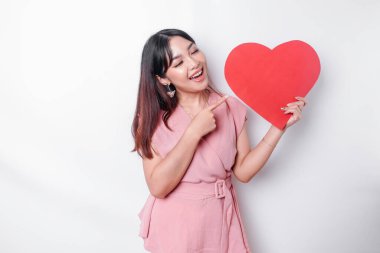 Excited Asian woman dressed in pink, pointing at the copy space beside her while holding a big red heart-shaped paper, isolated by white background