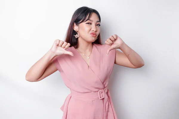 Disappointed Asian woman wearing pink blouse gives thumbs down hand gesture of disapproval, isolated by white background