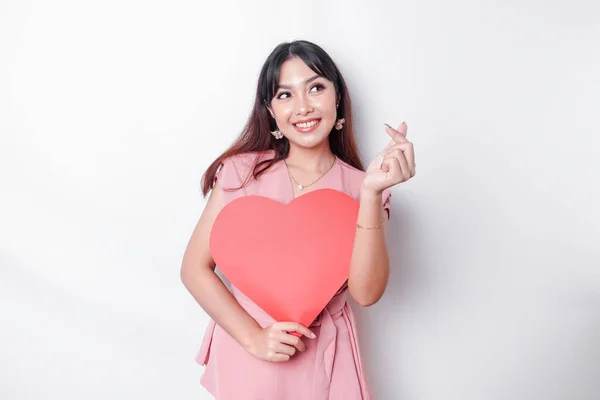 A happy young Asian woman wearing a pink blouse feels romantic shapes heart gesture expressing tender feelings and holding a red heart-shaped paper