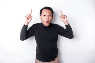 Shocked Asian man wearing black shirt pointing at the copy space on top of him, isolated by white background