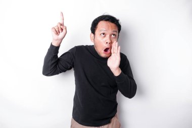 Shocked Asian man wearing black shirt pointing at the copy space on top of him, isolated by white background