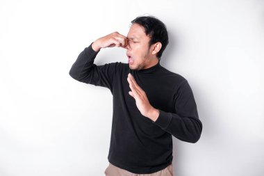 Young Asian man wearing t-shirt standing over isolated white background smelling something stinky and disgusting, intolerable smell, holding breath with fingers on nose. Bad smells concept.