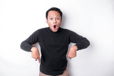Shocked Asian man wearing black shirt, pointing at the copy space below him, isolated by white background