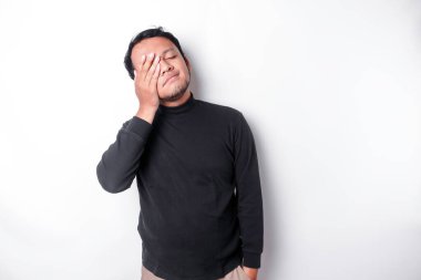 Portrait of sleepy attractive Asian man wearing a black shirt, feeling tired after night without sleep, yawning, covering opened mouth with palm