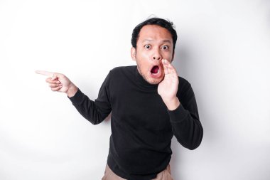 Shocked Asian man wearing black shirt pointing at the copy space beside him, isolated by white background