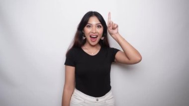 Excited Asian woman dressed in black shirt, pointing at the copy space on top of her, isolated by white background 