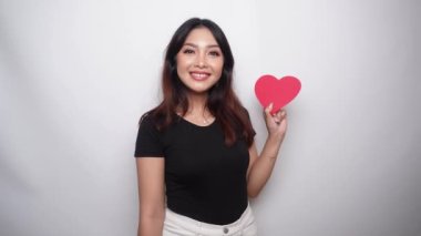 A happy young Asian woman wearing a black shirt feels romantic shapes heart gesture expressing tender feelings and holding a red heart shaped paper