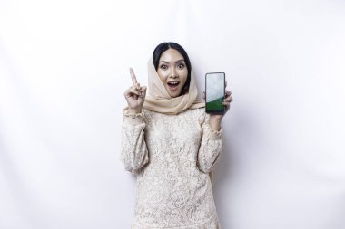 Excited Asian Muslim woman wearing hijab pointing at the copy space on top of her while showing her phone, isolated by white background