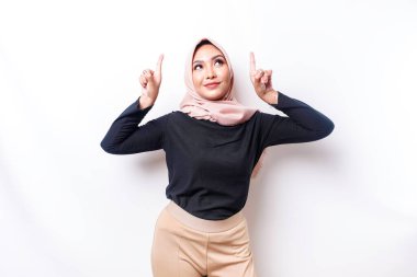 Excited Asian Muslim woman wearing hijab pointing at the copy space above her, isolated by white background