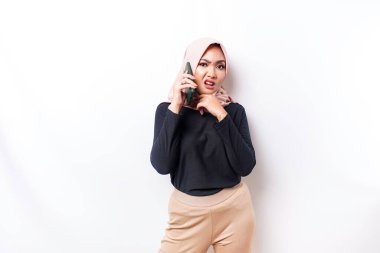 A thoughtful young Asian Muslim woman is wearing hijab and looks confused between choices while talking on her phone, isolated by a white background