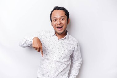 Excited Asian man wearing white shirt pointing at the copy space below him, isolated by white background