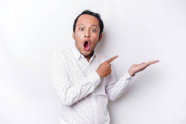 Shocked Asian man wearing white shirt pointing at the copy space beside him, isolated by white background