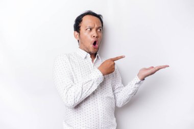 Shocked Asian man wearing white shirt pointing at the copy space beside him, isolated by white background