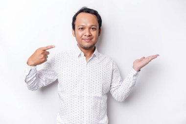 Excited Asian man wearing white shirt pointing at the copy space beside him, isolated by white background