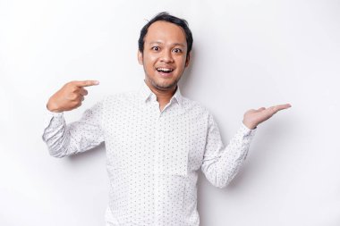Excited Asian man wearing white shirt pointing at the copy space beside him, isolated by white background