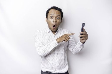 Shocked Asian man wearing a white shirt and holding his phone, isolated by white background