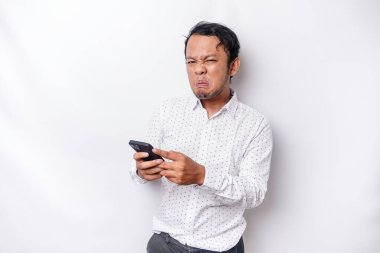 An angry young Asian man looks disgruntled wearing a white shirt irritated face expressions holding his phone