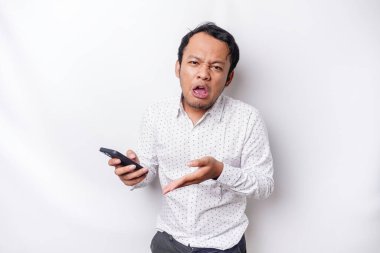 An angry young Asian man looks disgruntled wearing a white shirt irritated face expressions holding his phone