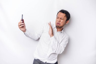 Young Asian man isolated on white background, looks depressed, face covered by arms frightened and nervous while holding his phone