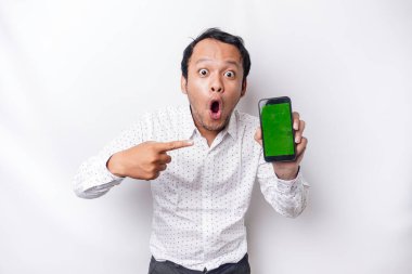 Shocked Asian man wearing a white shirt and showing green screen on his phone, isolated by white background