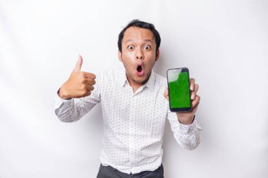 Shocked Asian man wearing a white shirt and showing green screen on his phone, isolated by white background