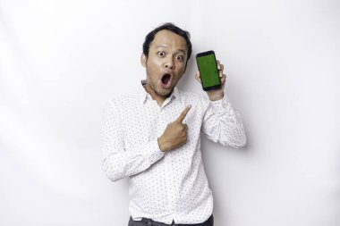 Shocked Asian man wearing a white shirt and showing green screen on his phone, isolated by white background