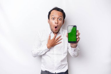 Shocked Asian man wearing a white shirt and showing green screen on his phone, isolated by white background
