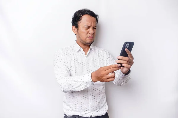An angry young Asian man looks disgruntled wearing a white shirt irritated face expressions holding his phone
