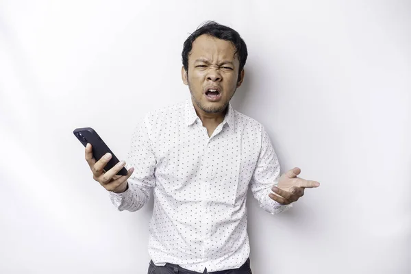 An angry young Asian man looks disgruntled wearing a white shirt irritated face expressions holding his phone