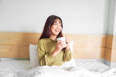 Süveter giyen, beyaz bir dairede yatakta çay içen ve gülümseyen güzel bir Asyalı kadın resmi.