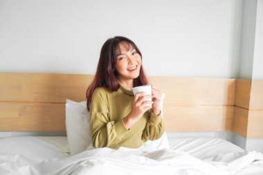 Süveter giyen, beyaz bir dairede yatakta çay içen ve gülümseyen güzel bir Asyalı kadın resmi.