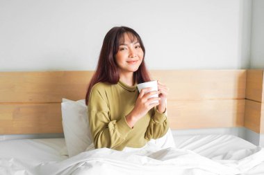 Süveter giyen, beyaz bir dairede yatakta çay içen ve gülümseyen güzel bir Asyalı kadın resmi.