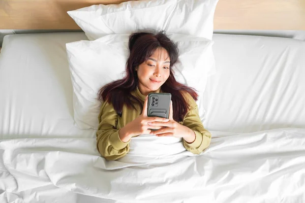 Улыбающаяся Молодая Азиатка Смеется Держа Телефон Лежа Кровати — стоковое фото