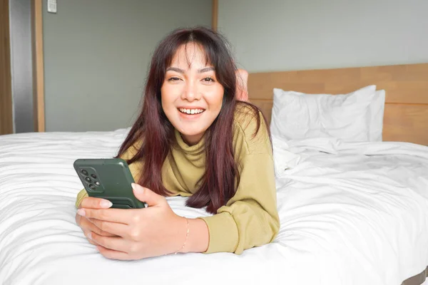 Smilende Asiatisk Kvinne Ler Mens Hun Holder Telefonen Ligger Senga – stockfoto