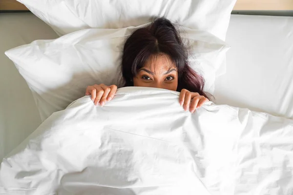 stock image A funny Asian woman lying in bed and hiding under a white blanket, looking at the camera with eyes full of joy.