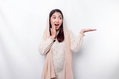 Shocked Asian Muslim woman pointing at the copy space on beside her, isolated by white background