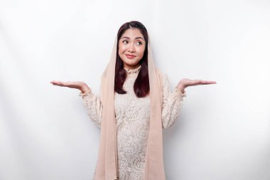 A thoughtful young Asian Muslim woman is wearing hijab and looks confused between choices, isolated by a white background