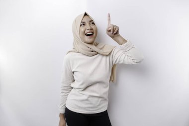 Excited Asian Muslim woman wearing hijab pointing at the copy space above her, isolated by white background