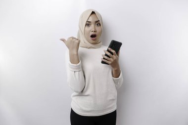 Shocked Asian Muslim woman wearing hijab pointing at the copy space beside her while holding her phone, isolated by white background