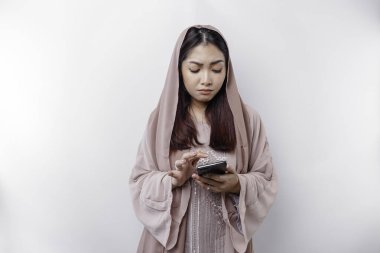 A dissatisfied young Asian Muslim woman looks disgruntled wearing a hijab irritated face expressions holding her phone