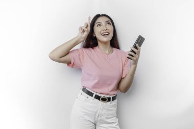 Pembe tişört giyen mutlu bir Asyalı kadının portresi. Telefonunu tutuyor ve üzerinde beyaz arka planla izole edilmiş fotokopi alanını işaret ediyor.