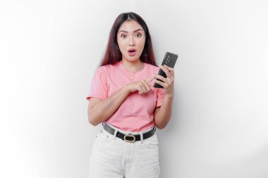 Shocked Asian woman dressed in pink and holding her phone, isolated by white background