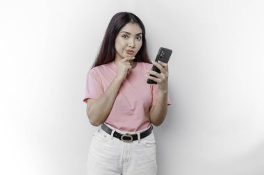 Pembe tişört giyen, çenesini ve telefonunu beyaz arka planda tutan düşünceli genç bir kadın.