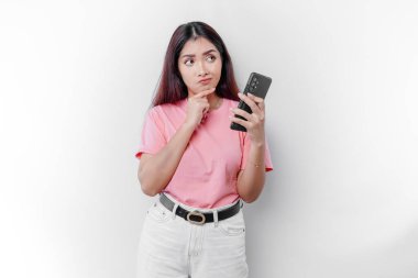 Pembe tişört giyen, çenesini ve telefonunu beyaz arka planda tutan düşünceli genç bir kadın.