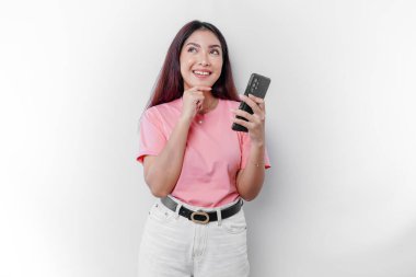 Pembe tişört giyen, çenesini ve telefonunu beyaz arka planda tutan düşünceli genç bir kadın.